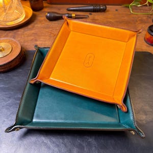 May include: Two square leather valet trays with raised, snapped corners. The larger tray is deep forest green, while the smaller tray is vibrant orange and features the embossed letters F and A stacked vertically inside an oval outline.