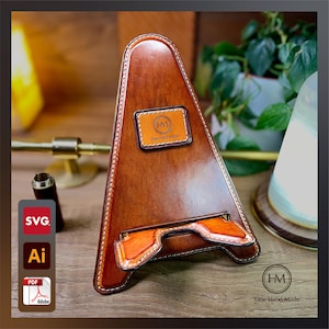 Pode incluir: Um suporte para telefone de couro marrom com um design costurado e um pequeno remendo de couro com o texto "HM One Hand Made".