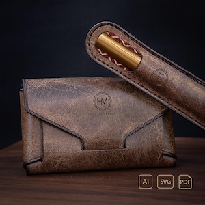 Może przedstawiać: Brązowy skórzany zestaw portfela i piórnika. Portfel ma zapięcie na klapę i napis "HM One Hand Made". Piórnik zawiera złoty długopis z czerwono-białym wzorem. Zestaw znajduje się na drewnianej powierzchni.