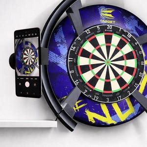 Könnte beinhalten: Eine kreisförmige Dartscheibe mit blau-gelbem Grafik-Hintergrund, montiert in einem schwarzen Ringrahmen. Ein Smartphone ist an der Seite des Rahmens befestigt und zeigt eine Live-Kameraansicht der Dartscheibe auf dem Bildschirm. Die Scheibe verfügt über standardmäßige nummerierte Segmente und das Winmau-Logo.
