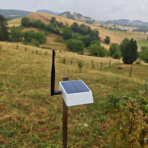Peut inclure: Un appareil solaire blanc avec une antenne noire monté sur un poteau en bois. L'unité dispose d'un panneau solaire rectangulaire sur sa surface supérieure et est placée à l'extérieur dans un champ herbeux avec des collines en arrière-plan.