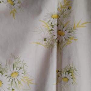 Puede incluir: Tela crema clara con un patrón repetido de margaritas. Las margaritas tienen pétalos blancos y centros amarillos, con hojas y tallos verdes. El fondo es de color crema claro.
