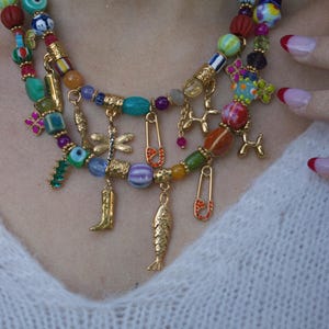 Puede incluir: Collar de cuentas multicolores de varias hebras. El collar presenta una variedad de cuentas de diferentes formas y colores, incluyendo rojo, azul, verde y dorado. Tiene colgantes dorados, incluyendo un pez, una bota y imperdibles.