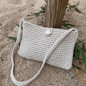 May include: A white crocheted bag with a shell accent. The bag has a long strap and is sitting on a sandy beach.
