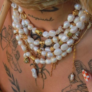Peut inclure: Collier de perles à plusieurs rangs avec des détails dorés. Le collier présente de grosses perles blanches irrégulières, des perles dorées et de petits ornements. Le design est superposé et orné, avec une esthétique vintage.