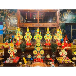 May include: A display of vibrant, decorative religious offerings. The offerings are arranged on a wooden table, featuring bright yellow, red, and blue elements. The scene is set in a room with detailed wall art and a wooden cabinet.