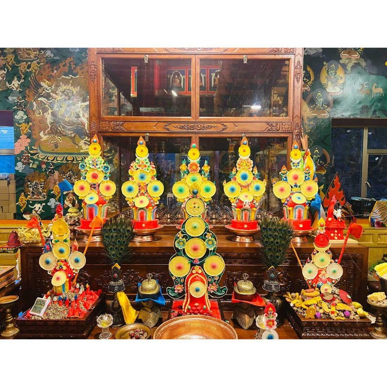 May include: A display of vibrant, decorative religious offerings. The offerings are arranged on a wooden table, featuring bright yellow, red, and blue elements. The scene is set in a room with detailed wall art and a wooden cabinet.