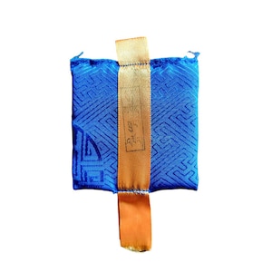 May include: A small, square blue fabric pouch with a geometric pattern. It has a gold-colored ribbon attached at the top and bottom, with text printed on the ribbon. The pouch is likely used for storage or as a decorative item.