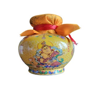 May include: A yellow ceramic jar with a rounded body and a decorative image of a seated figure. The jar is adorned with a yellow fabric cover tied with a red ribbon. The jar has a detailed, colorful design.
