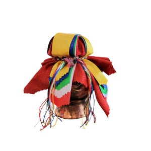May include: A decorative copper vessel is adorned with red, yellow, and multicolored ribbons and fabric. The vessel is wrapped in red fabric, with a yellow fabric top and colorful ribbons hanging down. The ribbons have blue, green, and red accents.