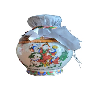 May include: A ceramic jar with a rounded body and a narrow neck, featuring a colorful illustration of a figure riding a mythical creature. The jar is adorned with intricate patterns and is covered with a white cloth tied at the top.