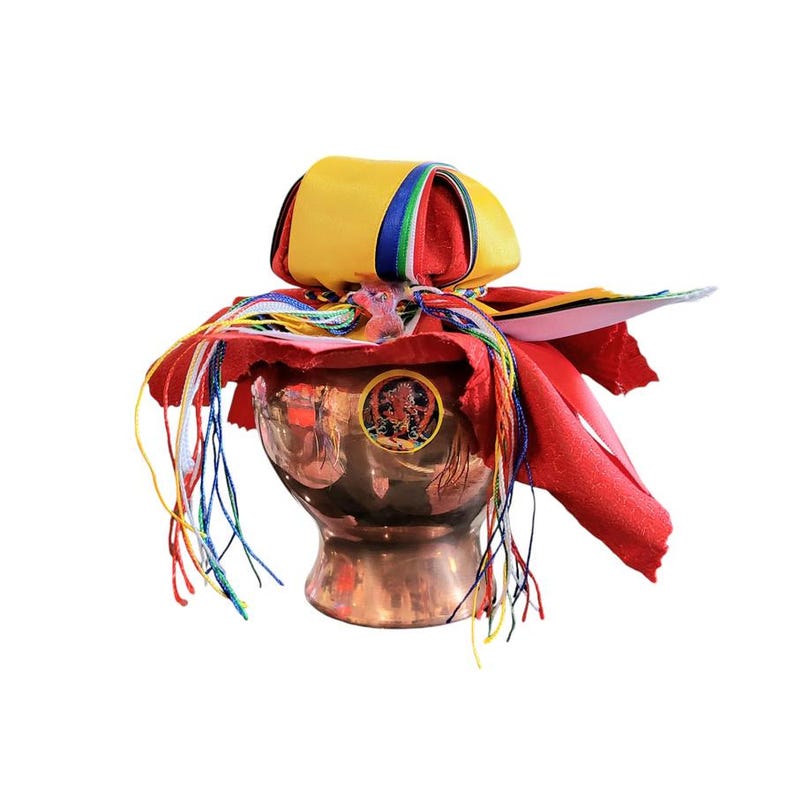 May include: A copper-colored ceremonial bowl with a decorative emblem, covered with a red cloth and a yellow and red fabric top. Colorful ribbons and tassels cascade from the top, adding a vibrant touch to this religious item.