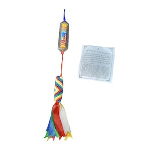 Puede incluir: Un colgante cilíndrico adornado con una sección trenzada colorida y una borla de cintas rojas, blancas, azules y verdes. El colgante está suspendido de un cordón azul. Un pequeño papel con texto está al lado del colgante.