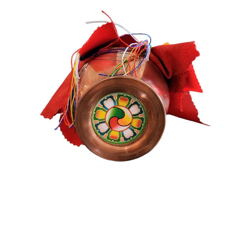 May include: A copper-colored Tibetan singing bowl with a colorful, floral-like design on the bottom. The bowl is partially covered with a red cloth and colorful strings. The bowl is likely used for meditation or spiritual practices.