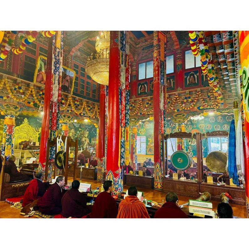 May include: Inside a Buddhist temple, featuring red pillars, elaborate decorations, and a large chandelier. Monks in maroon robes are seated on the floor, participating in a ceremony. The space is filled with colourful details and traditional artwork.