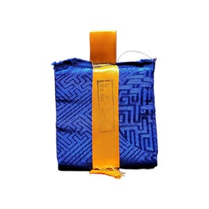 May include: A rectangular blue fabric pouch with a geometric pattern. It has a gold-colored ribbon and a small tassel at the bottom. The pouch has a small gold-colored tag with text.