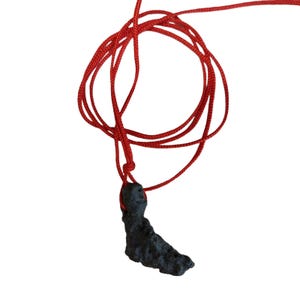 Könnte beinhalten: Ein schwarzer Anhänger mit unregelmäßiger Form hängt an einer roten Kordel. Die Kordel ist geschlungen und geknotet, wodurch eine Halskette entsteht. Der Anhänger scheint aus einem dunklen, möglicherweise steinartigen Material zu bestehen.