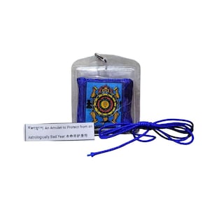 Puede incluir: Un amuleto azul con un diseño colorido dentro de una funda de plástico transparente. El amuleto está sujeto a un cordón azul. También se incluye una pequeña tarjeta blanca con texto en varios idiomas.