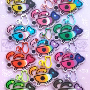 Puede incluir: Una colección de doce llaveros de acrílico con forma de peces estilizados con ojos grandes y pestañas largas. Cada llavero presenta una combinación de colores única y un cierre con forma de pez a juego, dispuestos en un patrón de cuadrícula.