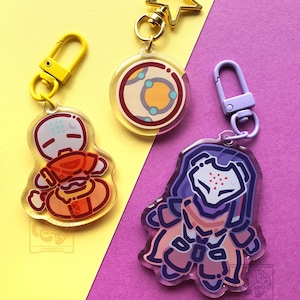 May include: Three acrylic keychains featuring stylised character illustrations. One keychain has a yellow clasp, one has a gold star-shaped clasp, and one has a light purple clasp. The designs include a round planet-like icon and two figures.