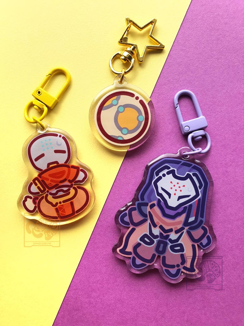 May include: Three acrylic keychains featuring stylized character illustrations. One keychain has a yellow clasp, one has a gold star-shaped clasp, and one has a light purple clasp. The designs include a round planet-like icon and two figures.
