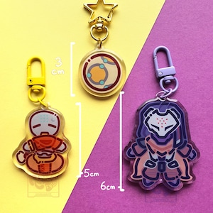 May include: Three acrylic keychains with metal clasps. One is a circular charm with a 3 cm diameter, one is a 5 cm tall orange character charm with a yellow clasp, and one is a 6 cm tall purple armored character charm with a purple clasp.