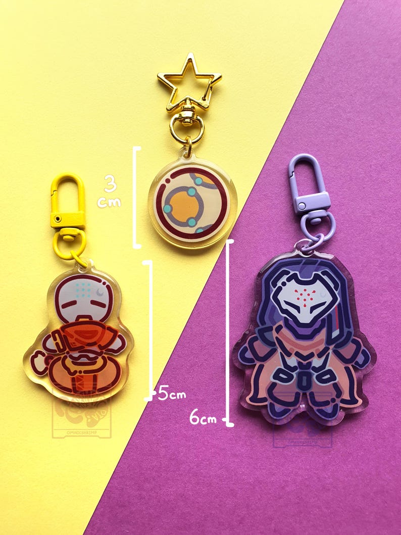 May include: Three acrylic keychains with metal clasps. One is a circular charm with a 3 cm diameter, one is a 5 cm tall orange character charm with a yellow clasp, and one is a 6 cm tall purple armored character charm with a purple clasp.