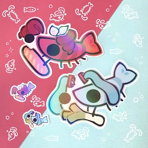 May include: Two die-cut stickers featuring stylized shrimp characters. One shrimp is pink and purple, holding a baguette. The other is blue and teal, holding a leek. Both have large dark eyes and are surrounded by smaller versions of the same designs.