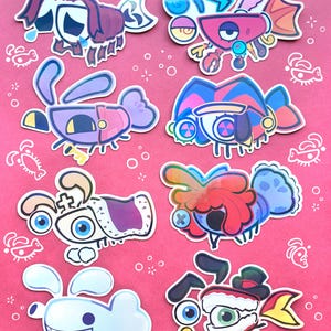 Op de afbeelding: Een verzameling van acht verschillende, kleurrijke stickers in cartoonstijl met gestileerde waterwezens met grote ogen, diverse accessoires en expressieve kenmerken, gerangschikt op een felroze achtergrond met kleine witte lijntekeningen.