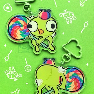 Puede incluir: Dos llaveros de acrílico con un personaje de dibujos animados verde de ojos grandes, que lleva una gorra de hélice colorida y sostiene una piruleta de remolino arcoíris. Cada llavero incluye un cierre verde claro en forma de nube.