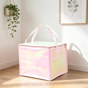 May include: A square-shaped insulated cooler bag with a shimmering, iridescent finish that reflects soft pink, yellow, and green hues. The bag features a zippered top closure and two white fabric handles attached to the top edges for carrying.