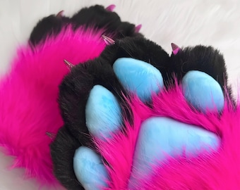 May include: A pair of furry paw gloves in vibrant colors. The gloves feature black fur with bright pink accents and light blue paw pads. The claws are a matching pink. The gloves are designed to resemble animal paws.