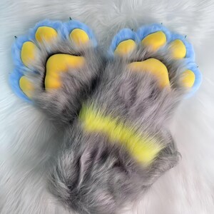 Puede incluir: Un par de guantes de pata peludos con pelaje gris, almohadillas azules claras y detalles amarillos. Las garras son negras. Los guantes están sobre una superficie blanca y esponjosa.