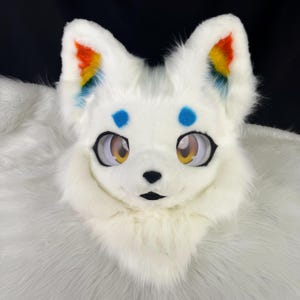 May include: A white faux fur animal mask featuring large, expressive eyes with yellow irises, a small black nose, and blue spots above the eyes. The inner ears are detailed with a vibrant rainbow gradient pattern, and the fur is thick and plush.