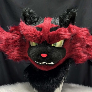 May include: A plush, furry mask in the shape of a cat-like creature. The mask features red fur with black accents, including the ears and facial markings. It has green eyes, a pink nose, and white teeth.