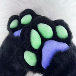 May include: Close-up of a pair of black furry paw gloves with green and purple paw pads. The gloves are made of a soft, plush material, and the paw pads are smooth and rounded. The background is white.