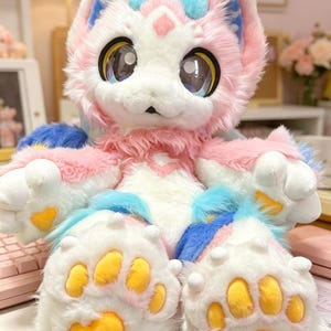 May include: A soft, plush creature in pastel pink, white, and blue. It has large eyes and yellow paw pads. The design is whimsical, possibly a toy or costume. The creature's fur is fluffy and textured, adding to its appeal.