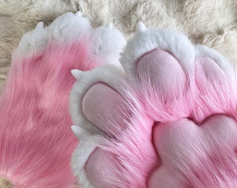 Puede incluir: Un par de patas peludas de color rosa y blanco con almohadillas de patas detalladas. Las patas son suaves y esponjosas, y las almohadillas son de color rosa claro.