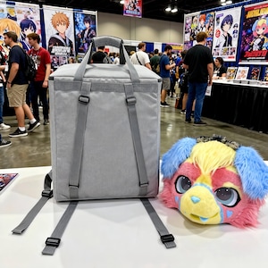 Puede incluir: Una mochila gris rectangular con correas ajustables y un asa superior está junto a una colorida cabeza de perro de peluche. La cabeza del perro presenta pelaje azul, amarillo, rosa y rojo, con ojos grandes y detalles en forma de corazón.