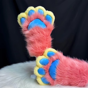 May include: A pair of furry paw-shaped costume feet in shades of pink, yellow, and blue. The paws have a plush, textured appearance with yellow pads and blue toe beans. The background is a dark fabric and white fur.