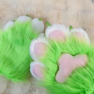 Puede incluir: Un par de patas peludas verdes y blancas con almohadillas rosadas. Las patas están hechas de tela suave y afelpada y están diseñadas para parecer patas de animales.