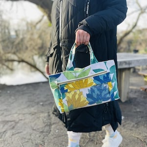 Könnte beinhalten: Eine Blumentasche mit weißem Hintergrund und blauen, grünen und gelben Blumen. Die Tasche hat eine Reißverschlusstasche und einen oberen Griff.