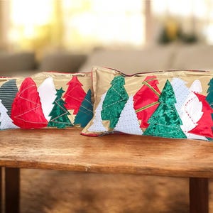 Könnte beinhalten: Zwei rechteckige Zierkissen mit goldenem Hintergrund und Weihnachtsbaum-Designs. Die Bäume sind in verschiedenen Farben gehalten, darunter Rot, Grün, Weiß und Blau. Die Kissen liegen auf einer Holzoberfläche.
