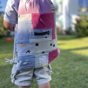 Könnte beinhalten: Ein Patchwork-Rucksack mit Kordelzugverschluss. Die Tasche hat eine Mischung aus rosa, blauen und grauen Stoffen mit einer kleinen Reißverschlusstasche. Das Design umfasst verschiedene Muster und Illustrationen, und die Tasche wird von einem Kind getragen.