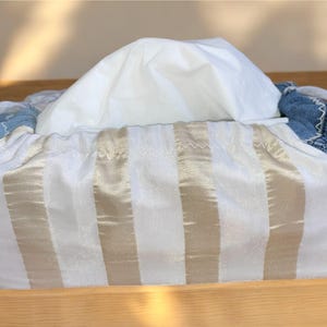 Könnte beinhalten: Ein rechteckiger Taschentuchbox-Bezug mit einer weiß-beige gestreiften Vorderseite. Die Seiten bestehen aus blauem Denim-Stoff. Weiße Taschentücher sind oben in der Box sichtbar.
