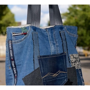 Könnte beinhalten: Eine blaue Jeans-Tragetasche mit mehreren Taschen und Patches. Die Tasche hat ein weißes Fleece-Futter und schwarze Tragegriffe. Die Tasche trägt den Text "TOMMY JEANS" wiederholt an der Seite.