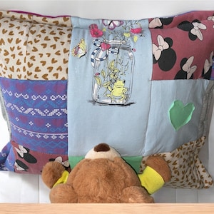 Könnte beinhalten: Ein Patchwork-Kissen mit verschiedenen Stoffen, darunter ein blauer Stoff mit einem Frosch-in-einem-Glas-Motiv, ein brauner Stoff mit einem Herzmuster und ein roter Stoff mit einem Mickey-Mouse-Schleifenmuster. Ein brauner Teddybär sitzt vor dem Kissen.