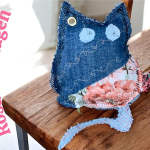 Könnte beinhalten: Ein handgefertigtes Katzenplüschtier aus Denim mit blauem Denim-Stoff und floral gemusterten Stoffakzenten. Die Katze hat zwei hellblaue kreisförmige Augen und eine goldene Öse. Der Text "Rosa mit Augen" ist sichtbar.