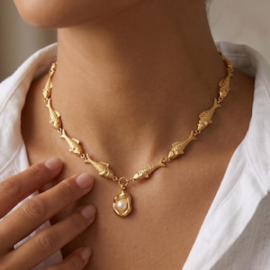 May include: Gold-toned necklace featuring a repeating fish design. The necklace has a pendant with a pearl. The jewelry is worn over a white button-down shirt.