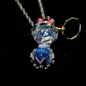 May include: A silver and gold necklace with a blue gem inside a cage-like pendant. The pendant is open and the gem is visible. The necklace has a small gold ring attached to it.